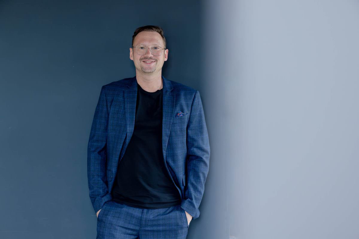 DJ Dennis steht fürs Portrait vor einer blauen Wand und hat die Hände in den Hosentaschen