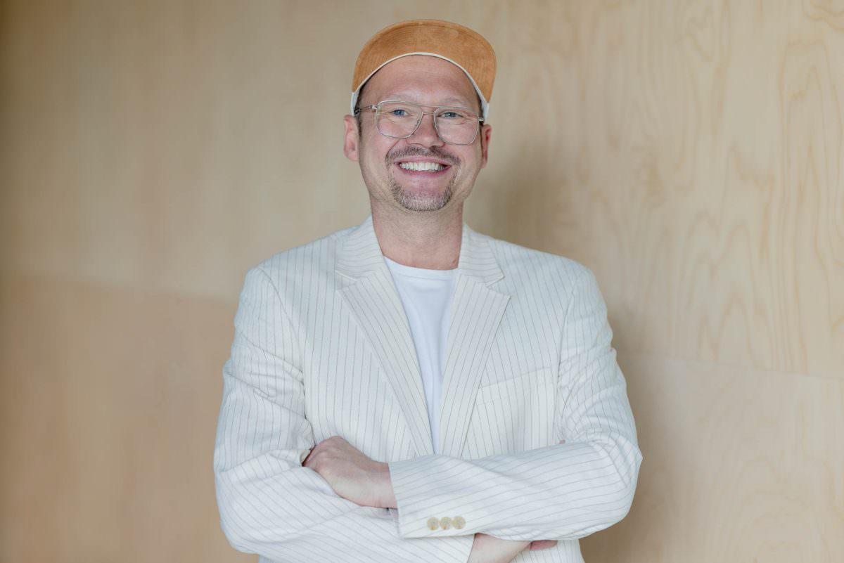 Halkörper Portrait von DJ Dennis, der mit verschränkten Armen vor einer hellen Holzwand steht