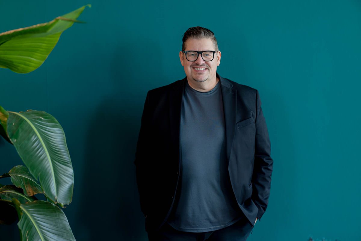 Half-body portrait of DJ Frank, standing in front of a dark green wall with his hands in his pockets.